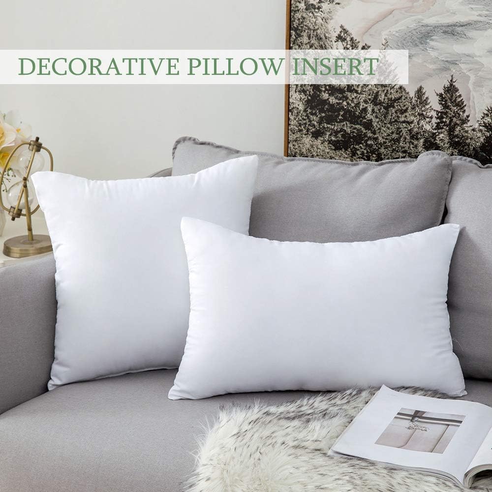 Two white decorative pillow inserts displayed on a gray sofa, with a text overlay reading 'DECORATIVE PILLOW INSERT' at the top. The setting includes a gray sofa, a white faux-fur throw, and a framed landscape print in the background.