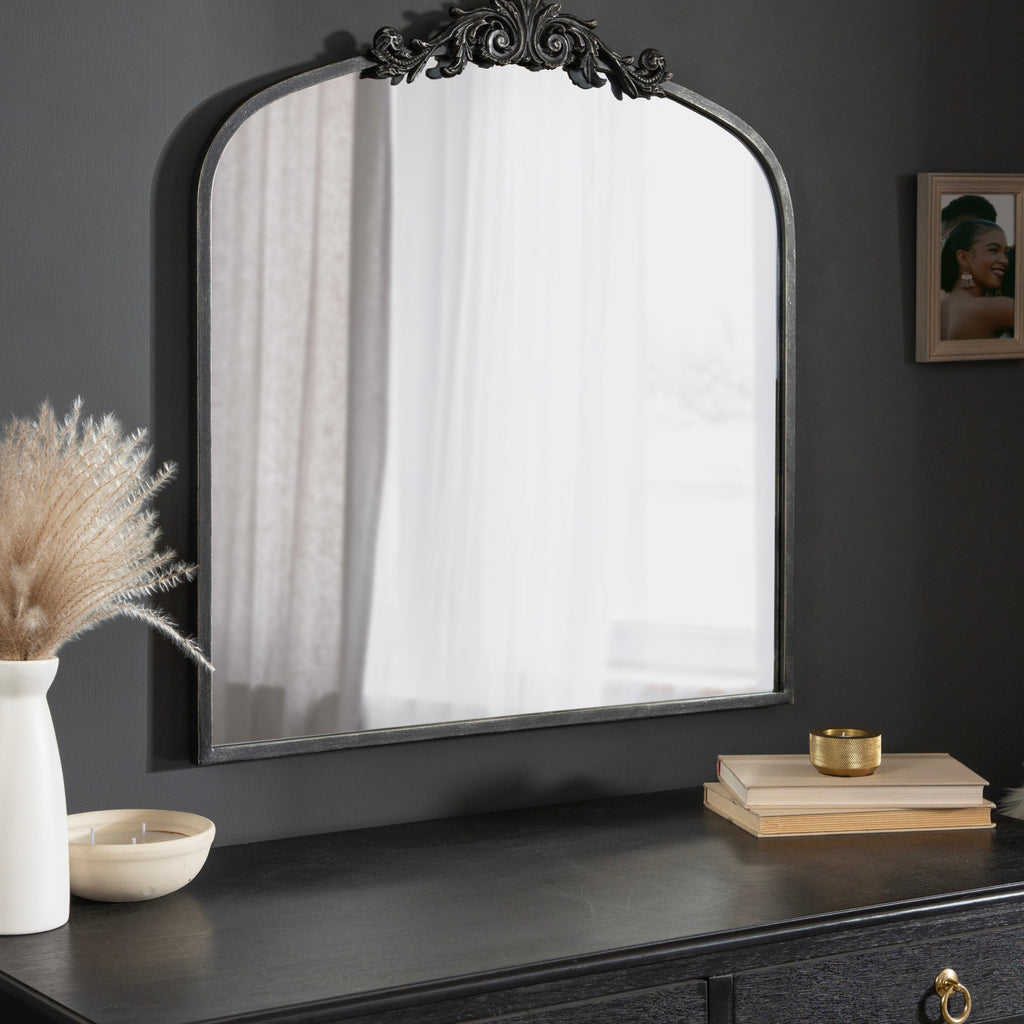 Decorative mirror on a dark wooden dresser with a vase and books in a dimly lit room.