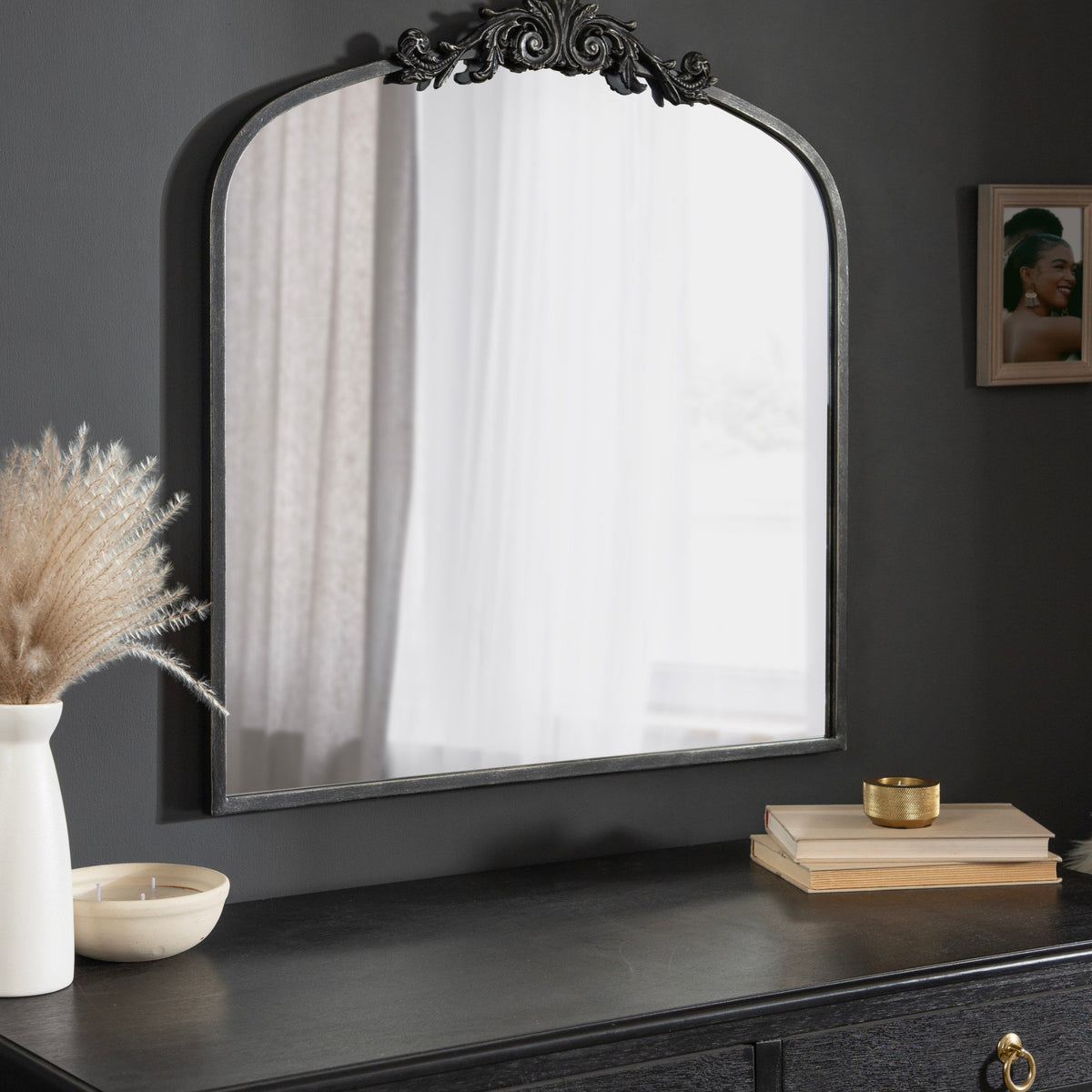 Decorative mirror on a dark wooden dresser with a vase and books in a dimly lit room.