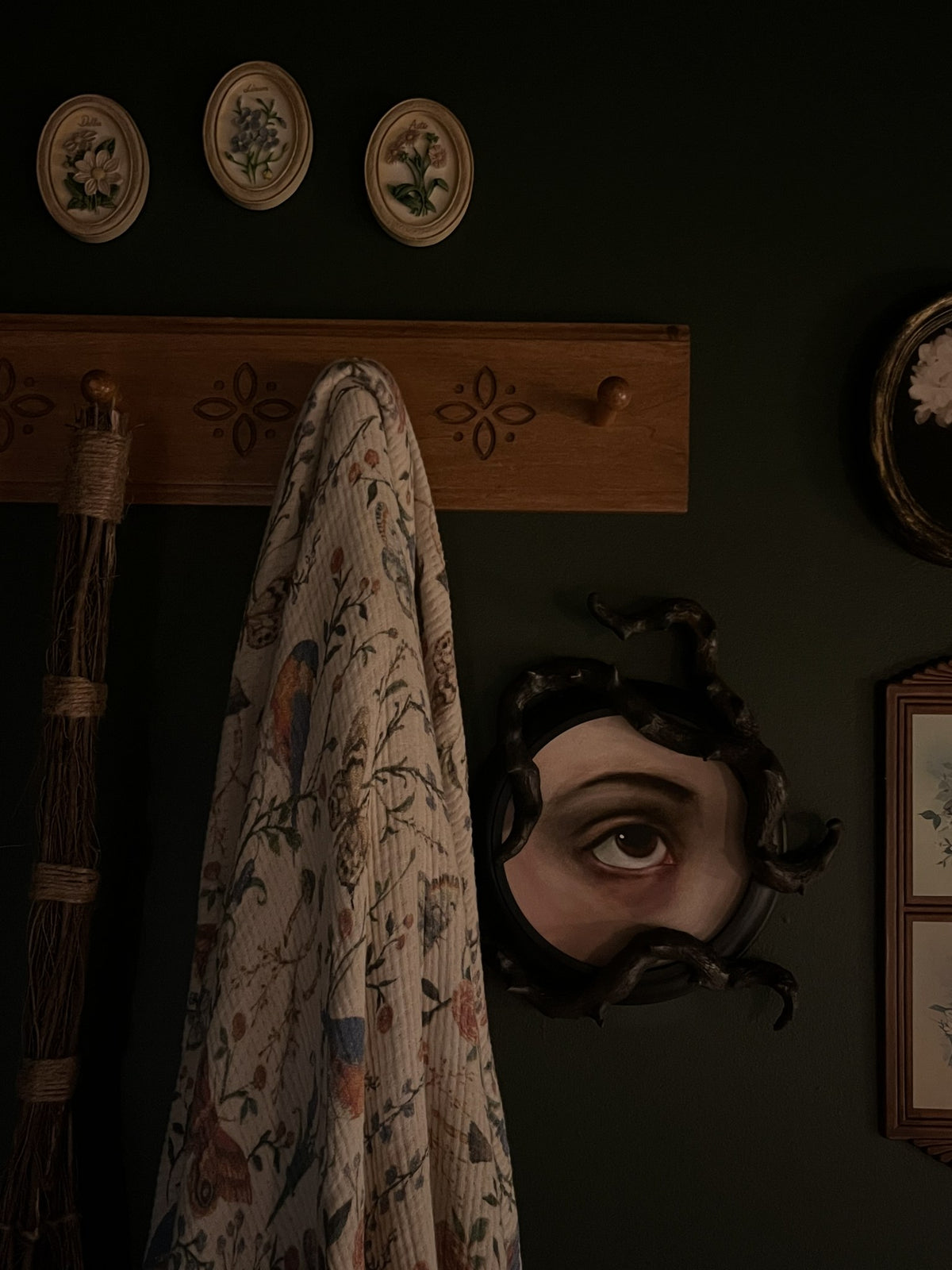 A vintage-style floral patterned throw blanket draped over a wooden wall hook in a dark, moody room. A decorative black eye mirror hangs on the wall, and other antique-style home decor items are visible in the background.
