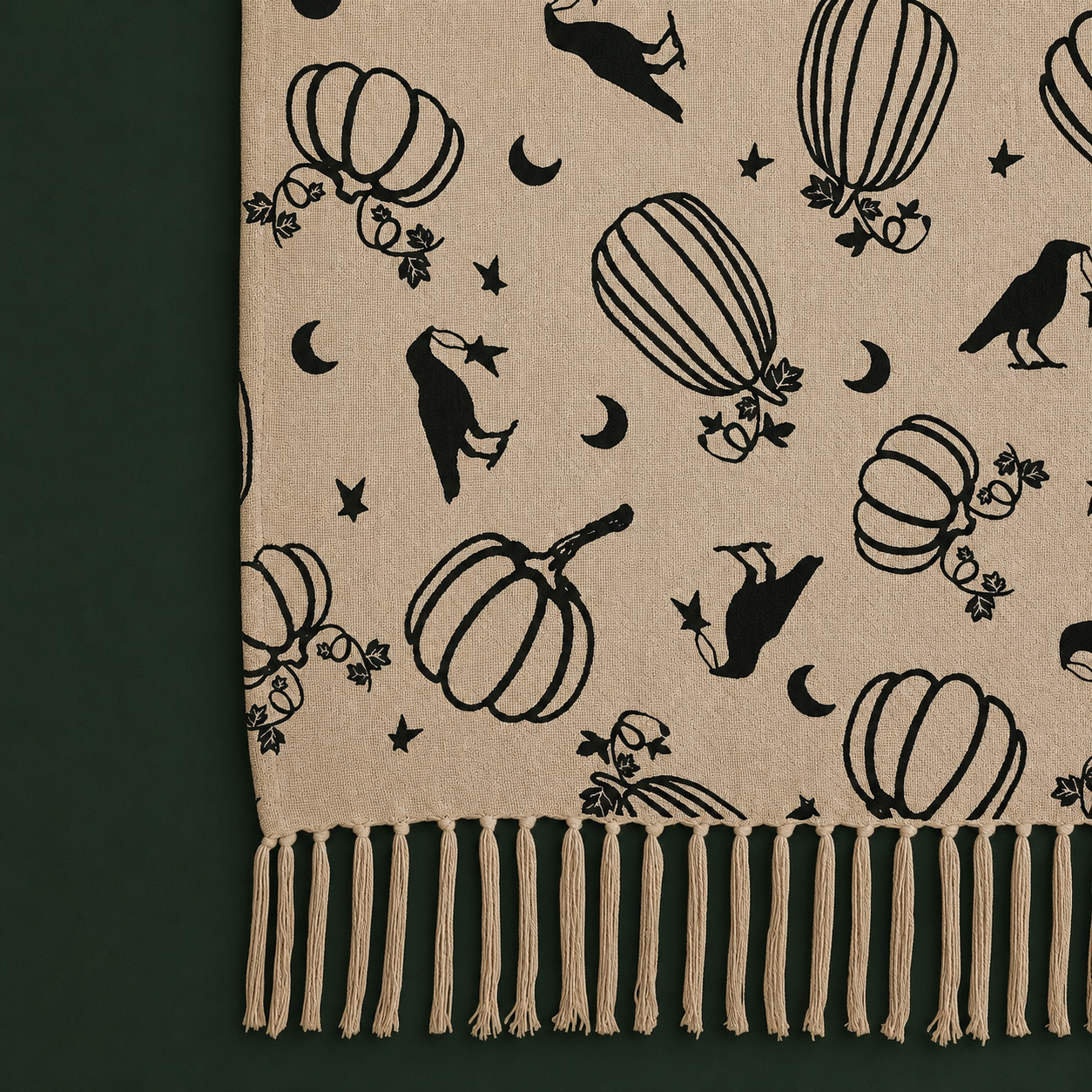 A close-up of a beige woven textile with black seasonal motifs, including pumpkins, crows, hot air balloons, stars, and crescent moons, finished with a fringed edge. The fabric is laid against a dark green background.