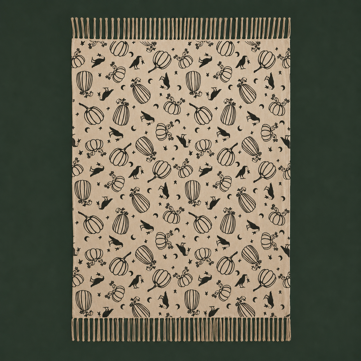 A rectangular woven throw blanket with fringe on all sides, featuring a black autumn-themed pattern of pumpkins, moons, stars, and birds on a warm beige background, set against a dark forest green backdrop.
