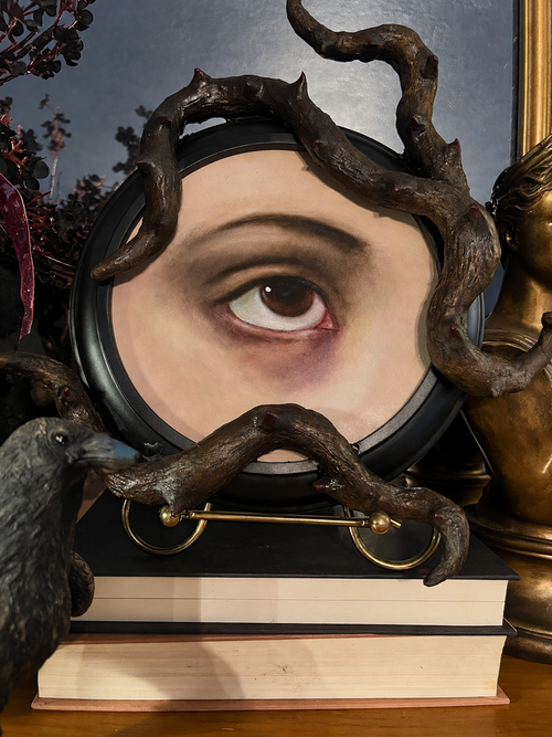 A decorative round mirror with a dark branch-like frame, shown on stacked books with a raven figurine at left and part of a bronze sculpture at right. The mirror reflects a painted human eye.