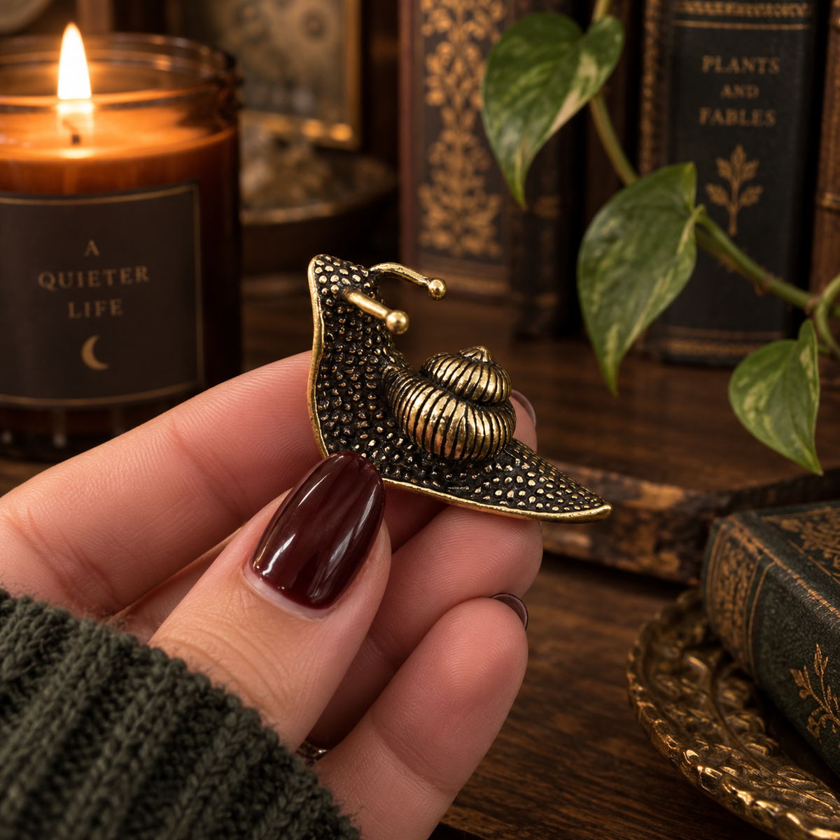 A hand with dark burgundy nail polish holds a small antique bronze snail charm against a cozy, vintage-themed background with a lit candle, books, and a green plant.