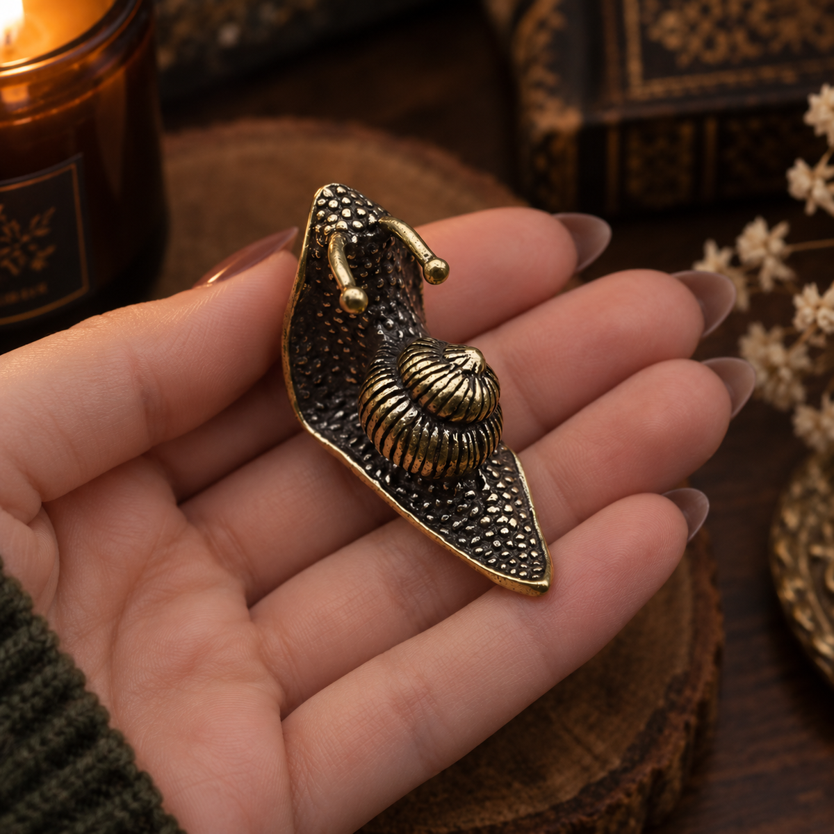 A close-up of a hand holding a vintage-style gold and bronze snail shell brooch against a warm, rustic background with a candle and books.