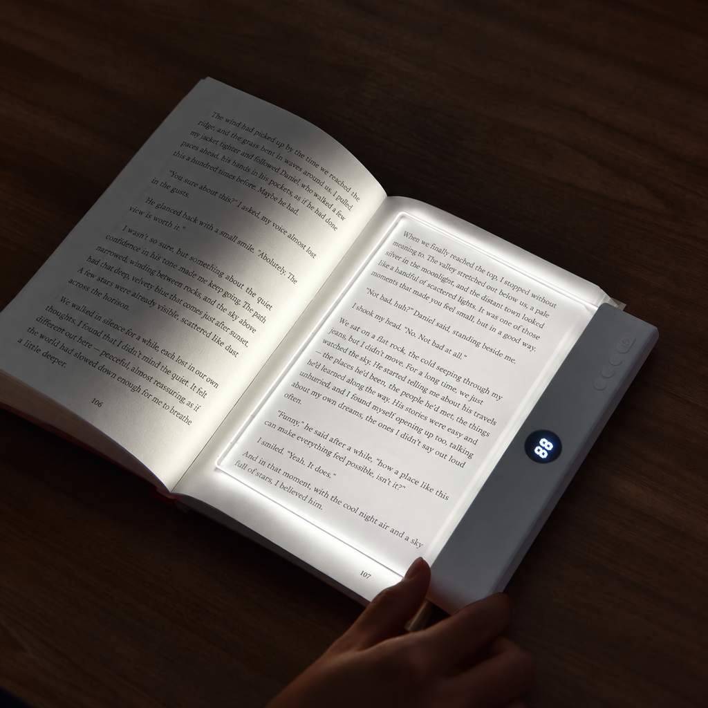 A hand holding an open white electronic book reader on a dark wooden table, showing illuminated pages with a small blue screen displaying text.