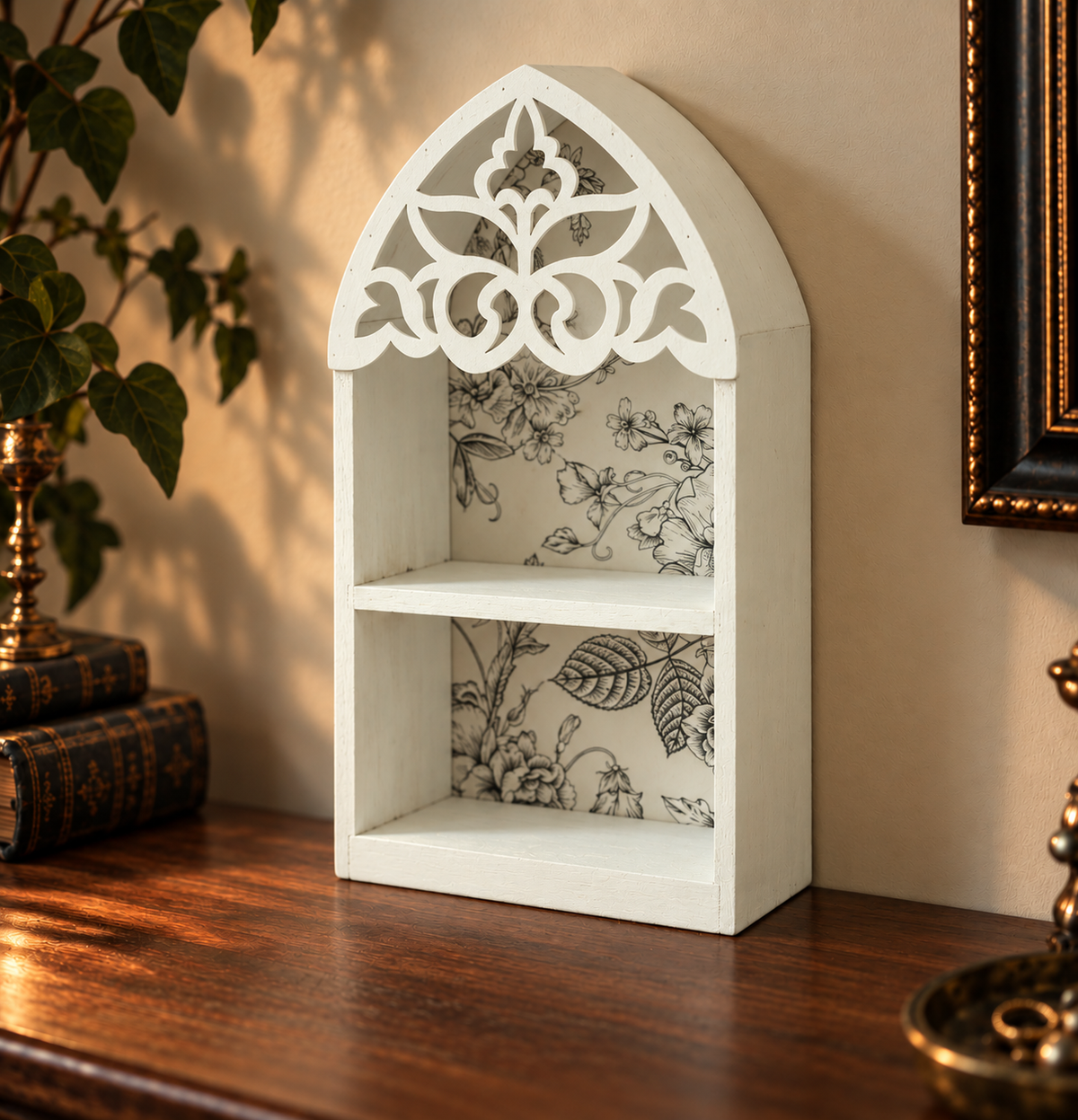 A small white arched wall shelf with ornate cutout detailing at the top and two open shelves, shown on a wooden table against a beige wall with decorative plants and framed art nearby.