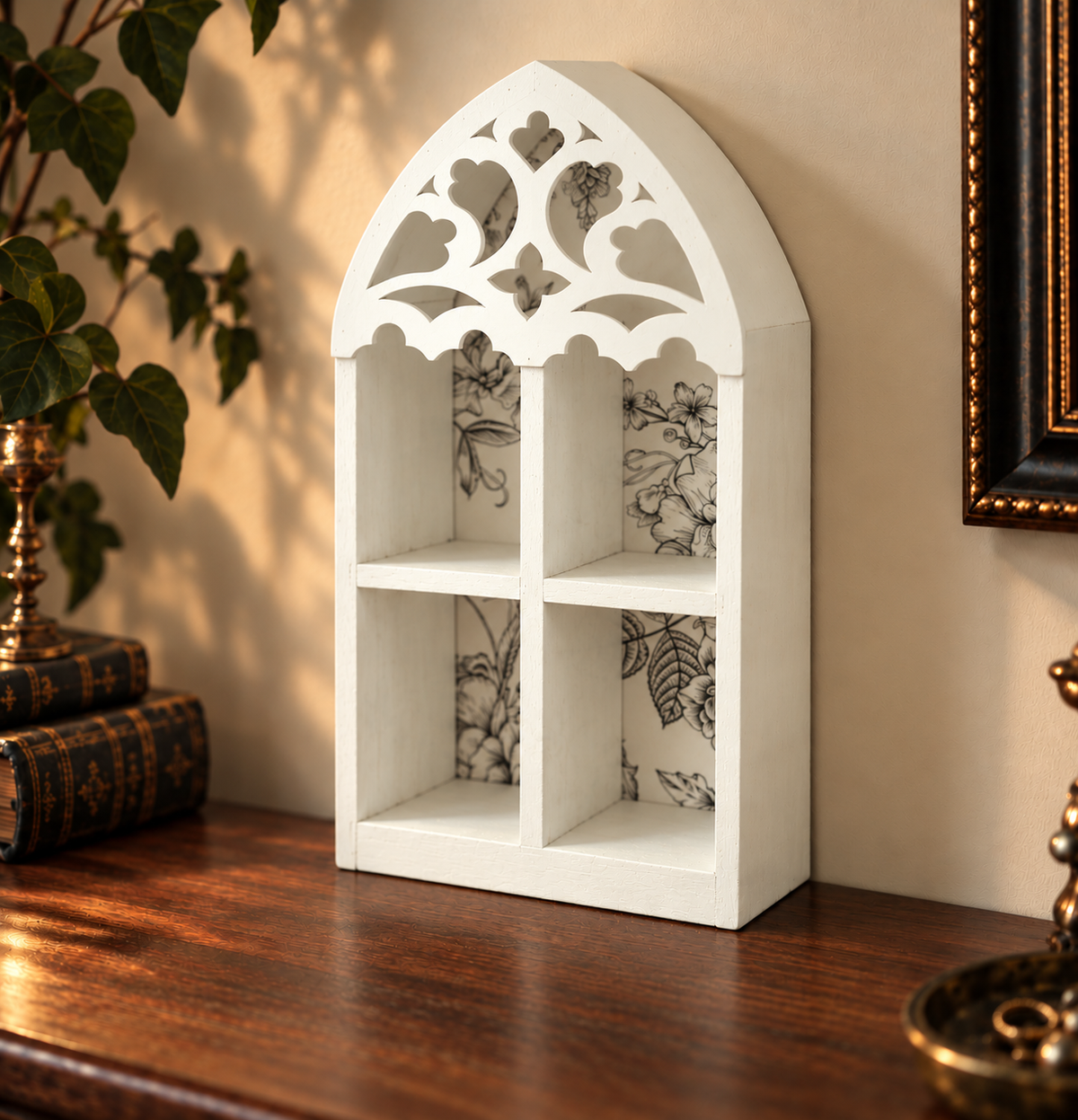 A white arched wall shelf with four compartments and decorative cutout detailing on the top, styled on a wooden table against a beige wall with books and a plant nearby.