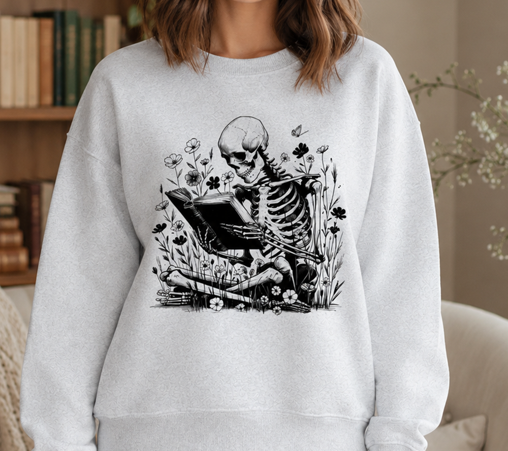 A woman wearing a light gray crewneck sweatshirt with a black graphic of a skeleton reading a book surrounded by wildflowers. The image is set in a cozy indoor environment with a bookshelf in the background.