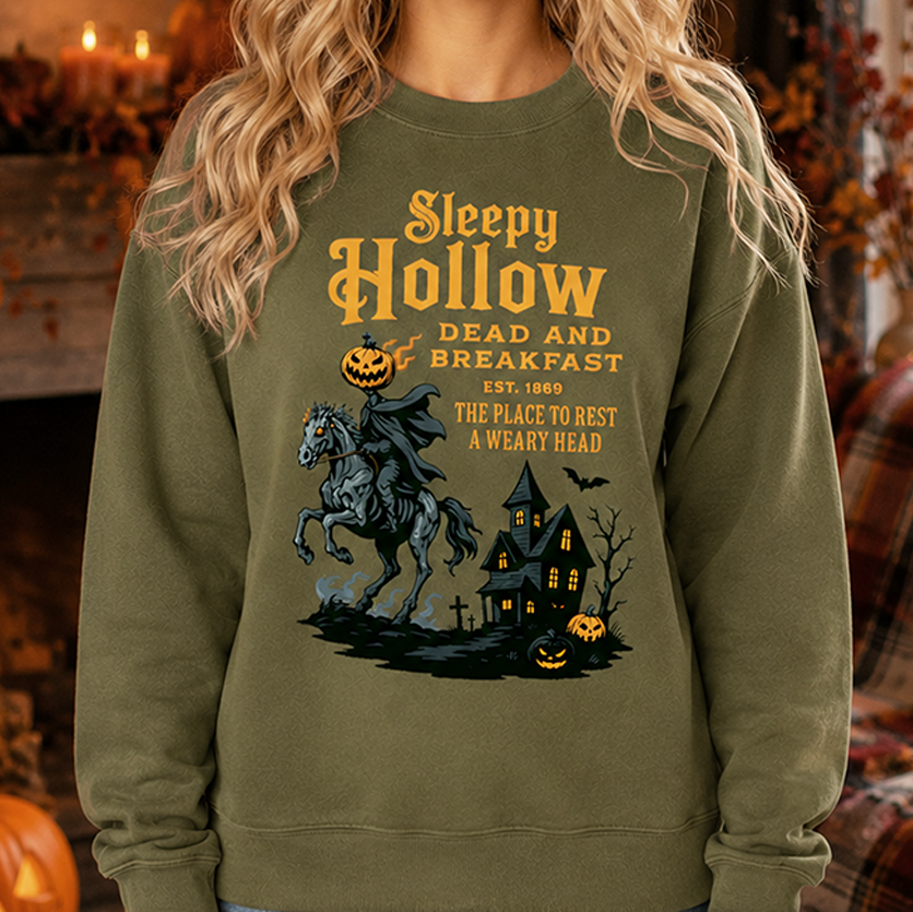 A woman wearing an olive green crewneck sweatshirt with a 'Sleepy Hollow' Halloween graphic, posed in a cozy, warmly lit holiday home setting.