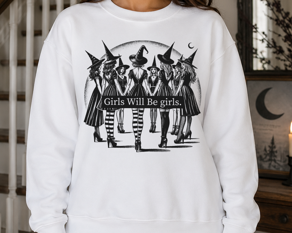 A woman wearing a white crewneck sweatshirt with a black graphic of five witches carrying a banner that reads 'Girls Will Be girls.' The image is taken indoors in a home setting with a staircase and framed art in the background.