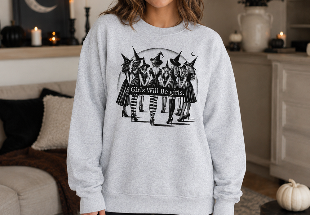 A woman wearing a light gray crewneck sweatshirt with a black graphic of witches and the text 'Girls Will Be girls.' in a cozy, decorated living room setting.
