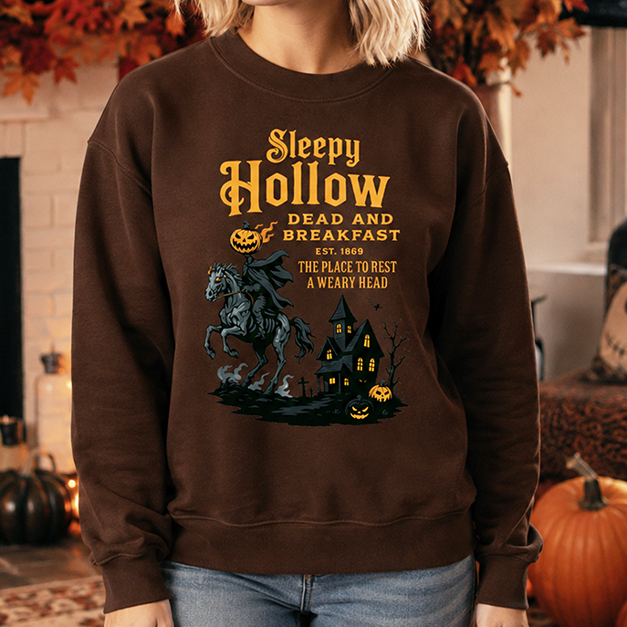 A woman wearing a brown crewneck sweatshirt with a 'Sleepy Hollow' graphic print, standing in a cozy, decorated Halloween-themed living room.