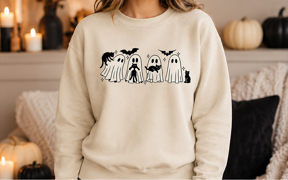 A woman wearing a cream-colored crewneck sweatshirt with a black graphic of five cute ghosts wearing Halloween costumes, posed in a cozy, warmly lit living room setting.