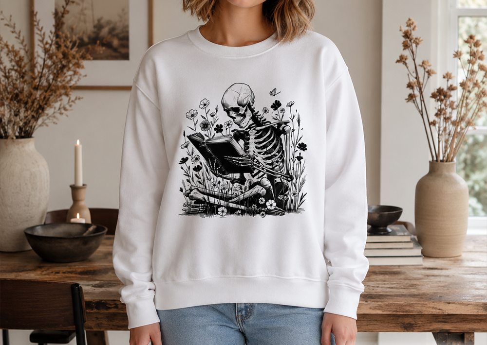 A woman wearing a white crewneck sweatshirt with a black graphic of a skeleton reading a book surrounded by flowers, standing in a styled room with a wooden table and vases.
