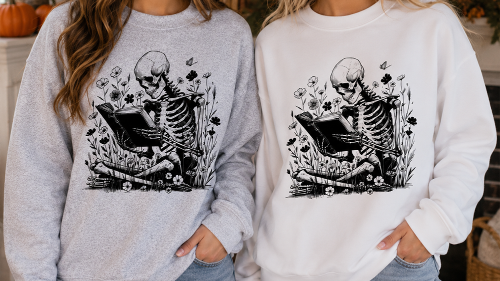 Two women standing side by side wearing matching crewneck sweatshirts with a black skeleton reading graphic. The woman on the left wears a light heather gray sweatshirt, and the woman on the right wears a white sweatshirt.