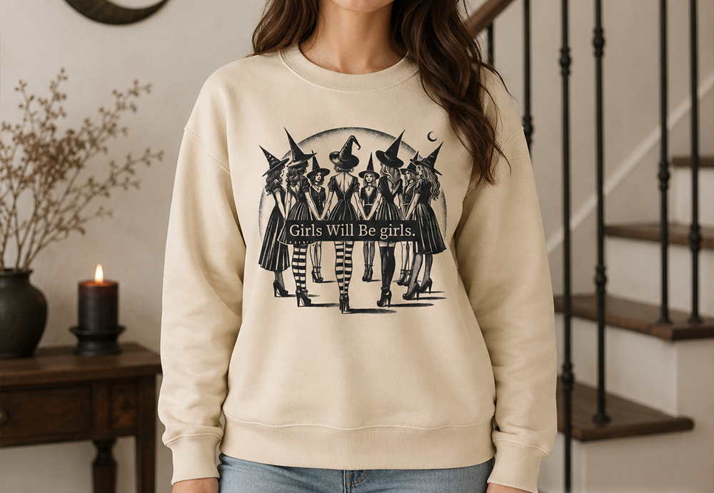 A woman wearing a cream-colored crewneck sweatshirt with a black graphic of five witches and the text 'Girls Will Be girls'. She is standing in a home interior with a staircase in the background.