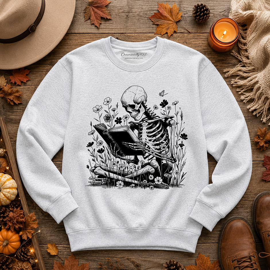 A light gray crewneck sweatshirt featuring a black and white graphic of a skeleton playing a violin in a field of flowers. The sweatshirt is displayed in a flat-lay arrangement on a rustic wooden surface, surrounded by autumn-themed props including a hat, a lit candle, leaves, and a pair of brown boots.