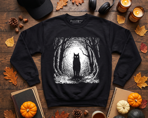 A black crewneck sweatshirt laid flat on a wooden surface, featuring a white line-art graphic of a cat standing in a forest. The sweatshirt is styled with autumn-themed props including fall leaves, candles, a black baseball cap, headphones, notebooks, pumpkins, pinecones, and a small cup.