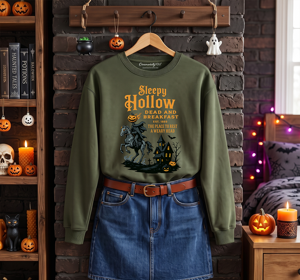 An olive green long-sleeve crewneck sweatshirt with a Halloween graphic print, styled on a wooden hanger over a blue denim skirt. The scene is set in a cozy, decorated bedroom with a brick wall background, featuring shelves of pumpkins, a bed with fairy lights, and a warm candlelit atmosphere.