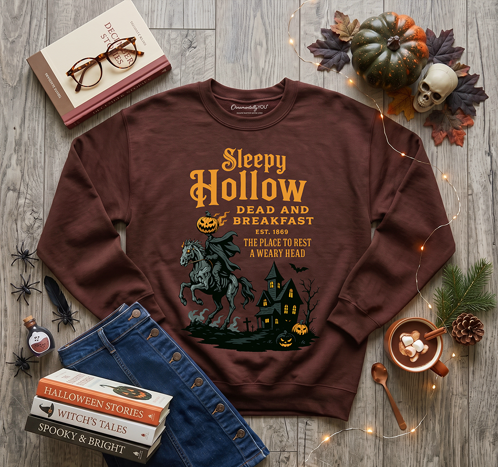 A flat-lay of a brown crewneck sweatshirt featuring a Halloween graphic with the text 'Sleepy Hollow Dead and Breakfast' and an illustration of a ghost horse, surrounded by themed props like books, glasses, a skull, and autumn leaves on a gray wood background.