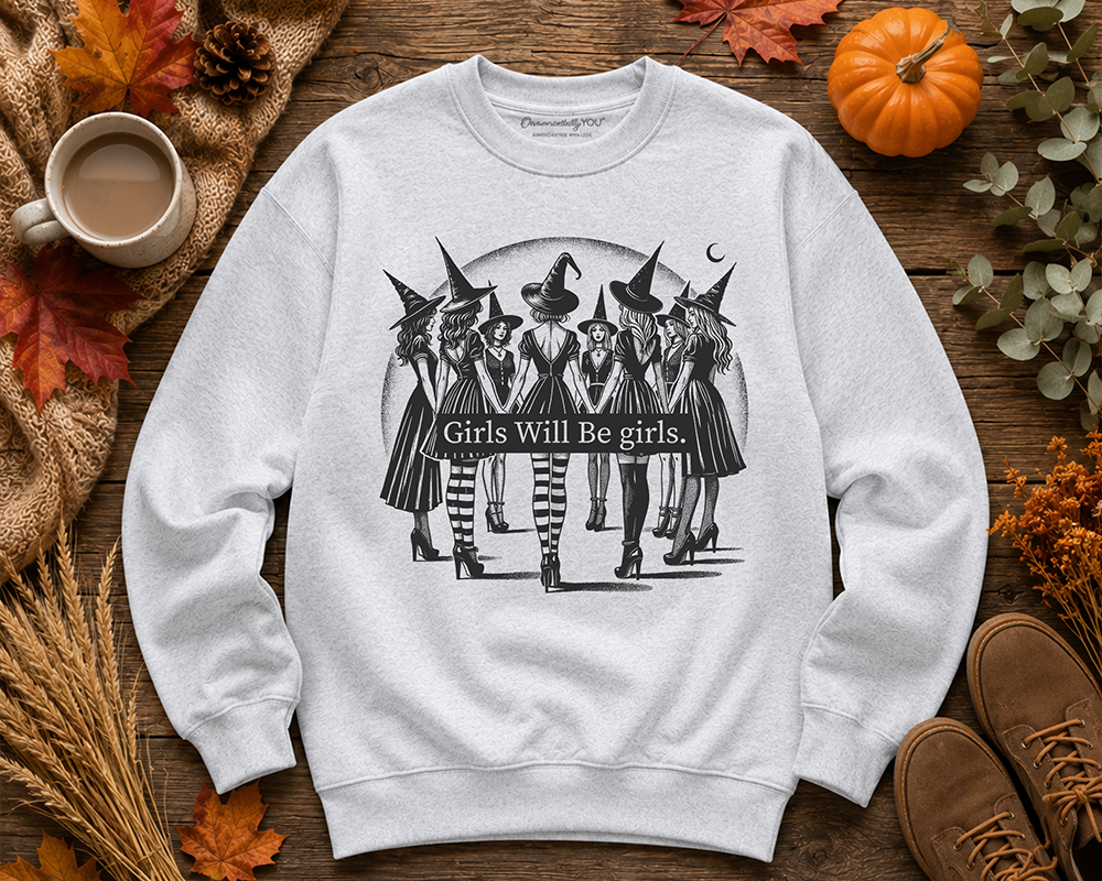 A light gray crewneck sweatshirt with a black graphic of five witches and the text 'Girls Will Be girls.' laid flat on a rustic wooden surface surrounded by autumn props including a pumpkin, leaves, a mug, and boots.