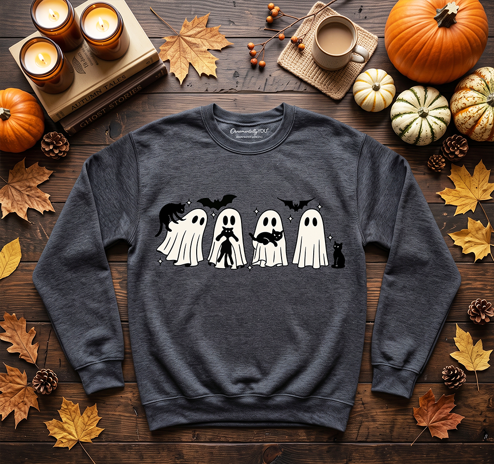 A dark heather gray crewneck sweatshirt with a Halloween graphic of four cute ghosts, styled in a flat-lay on a rustic wooden table surrounded by autumn leaves, pumpkins, candles, and a coffee mug.