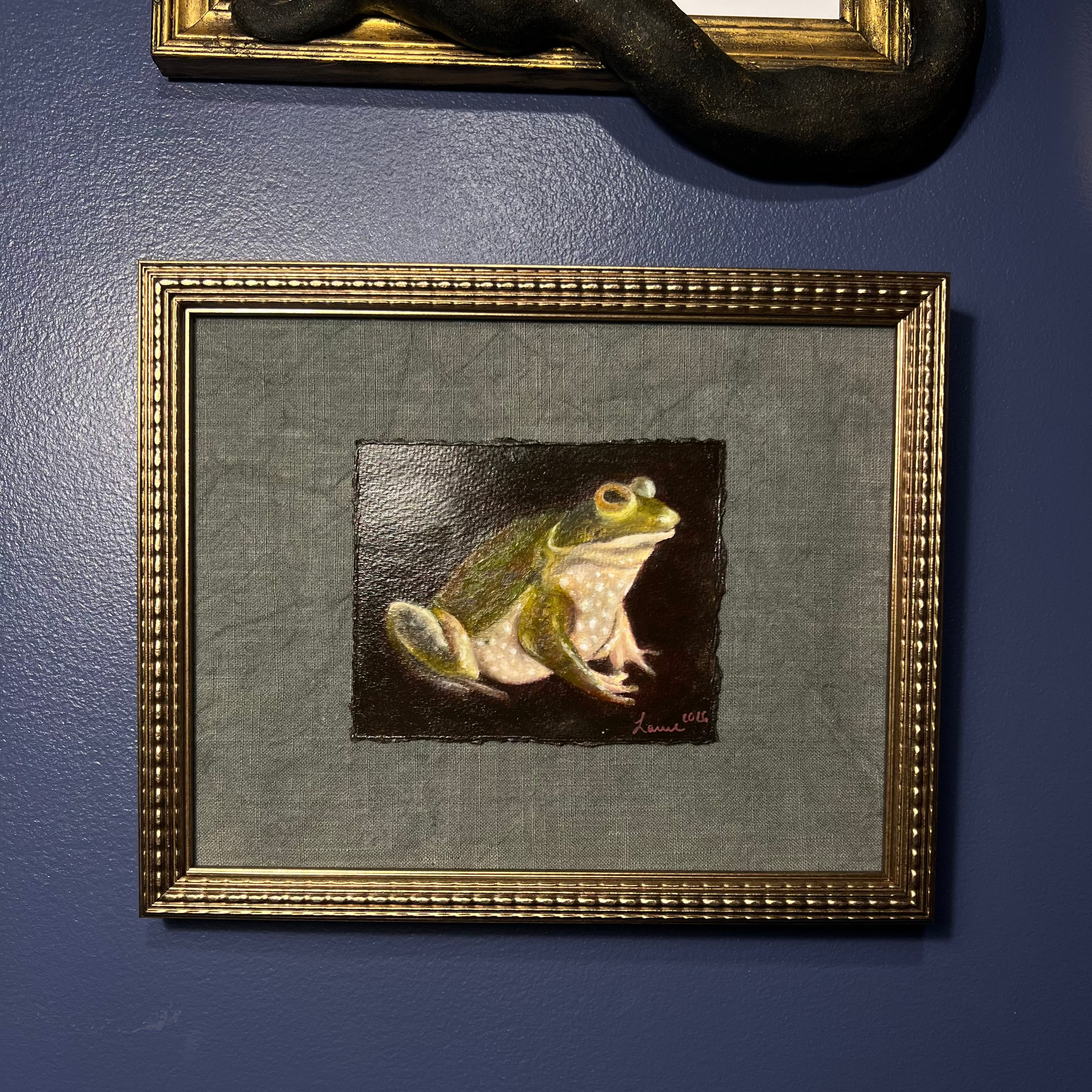 A framed painting of a green and yellow frog on a dark background, mounted on a textured blue wall. The artwork is enclosed in a gold-toned frame with a gray mat.