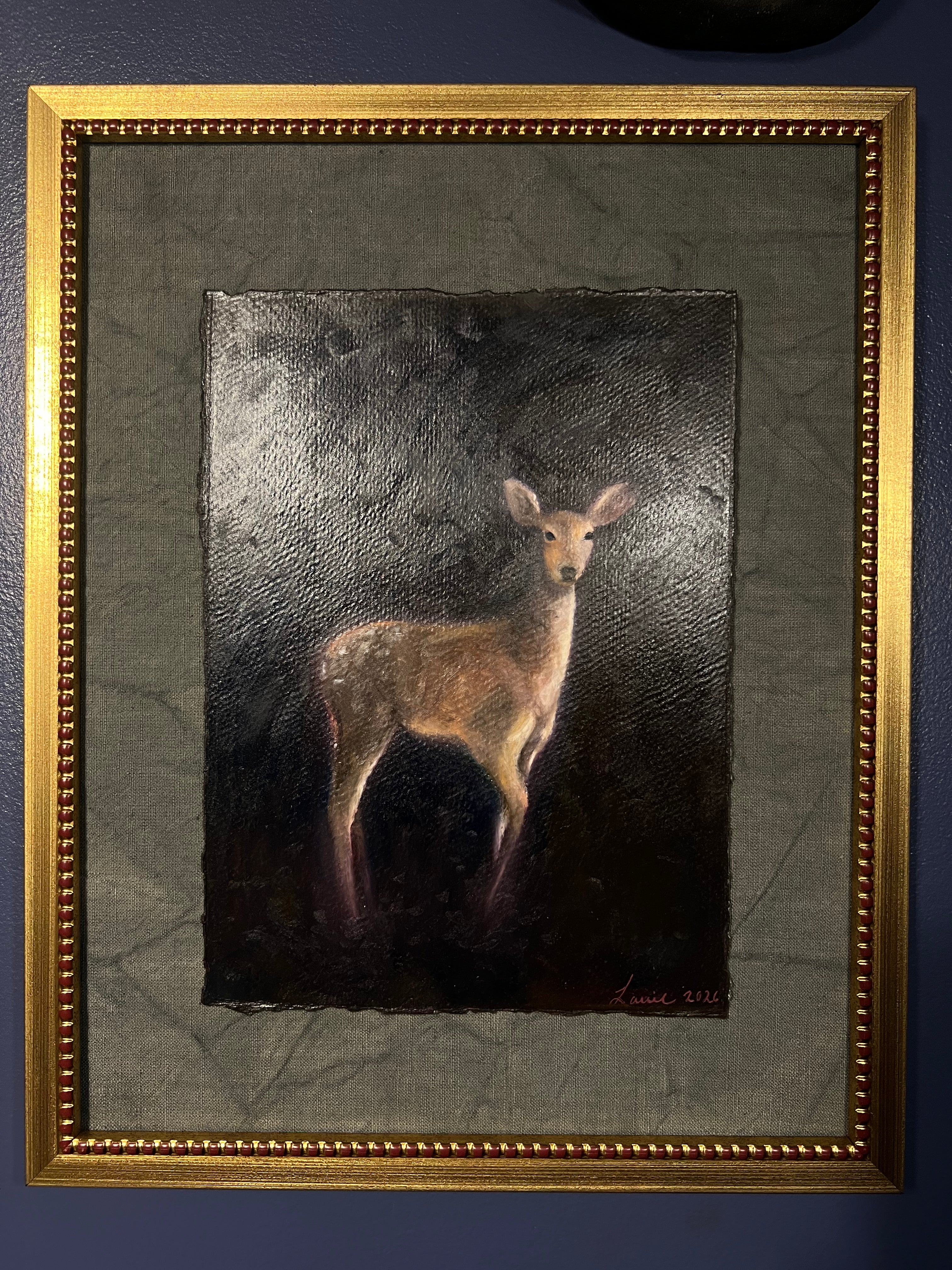 A framed painting of a deer standing against a dark textured background, enclosed in a wide gray mat and a decorative gold frame, hung on a dark blue wall.