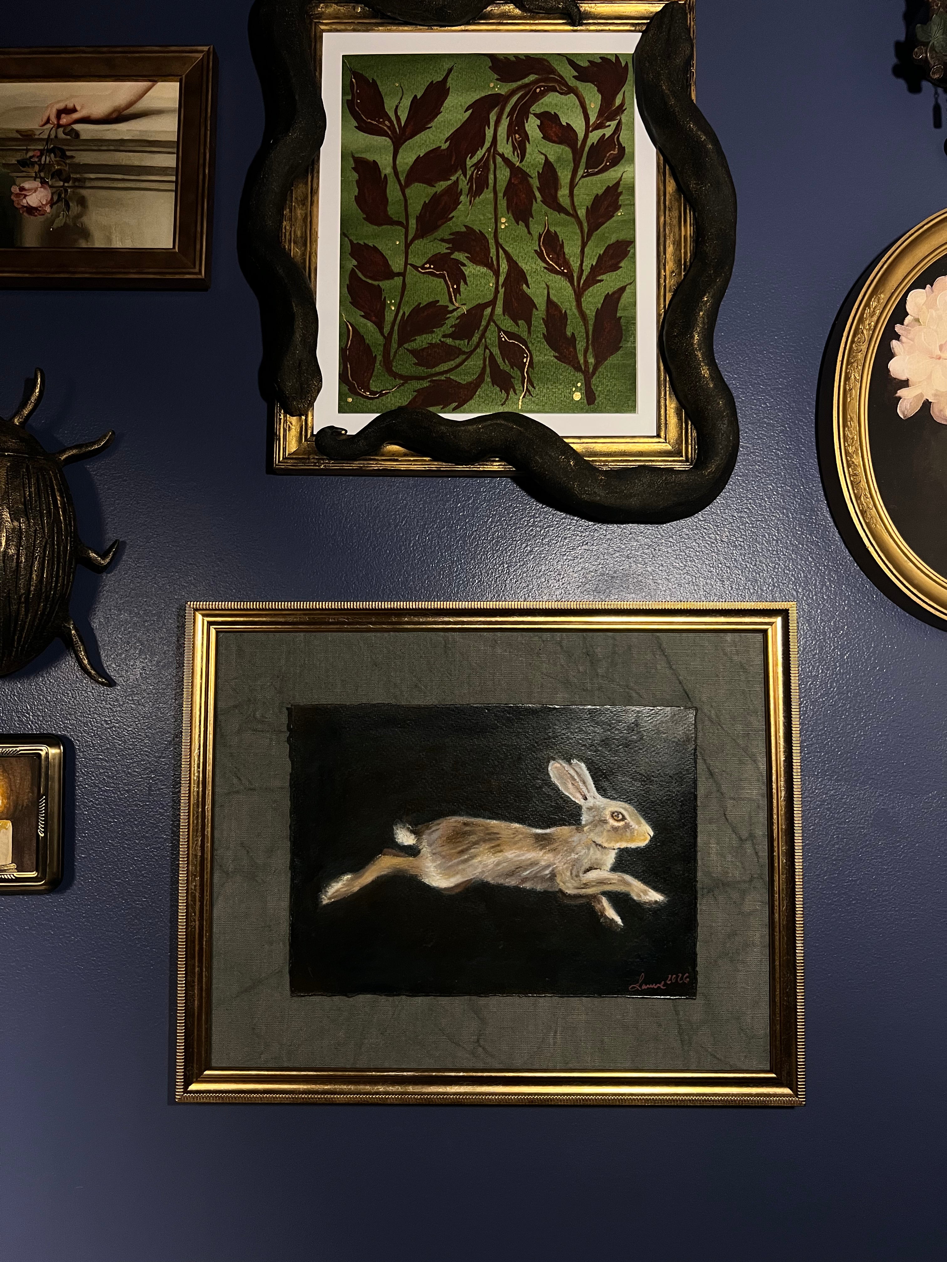 A framed art print depicting a leaping rabbit on a dark background, mounted on a dark blue wall alongside other decorative frames and objects.