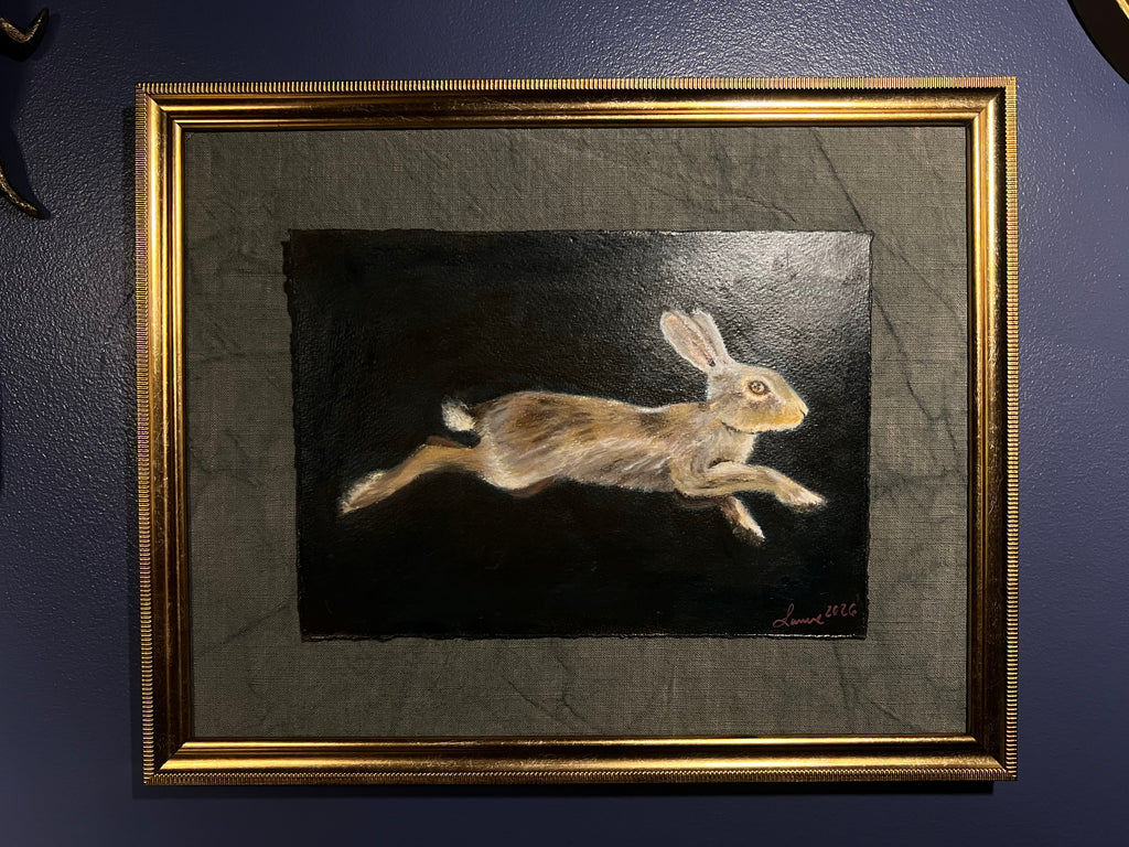 A framed painting of a leaping brown hare on a dark background, mounted on a textured gray mat within a decorative gold frame, displayed on a dark blue wall.