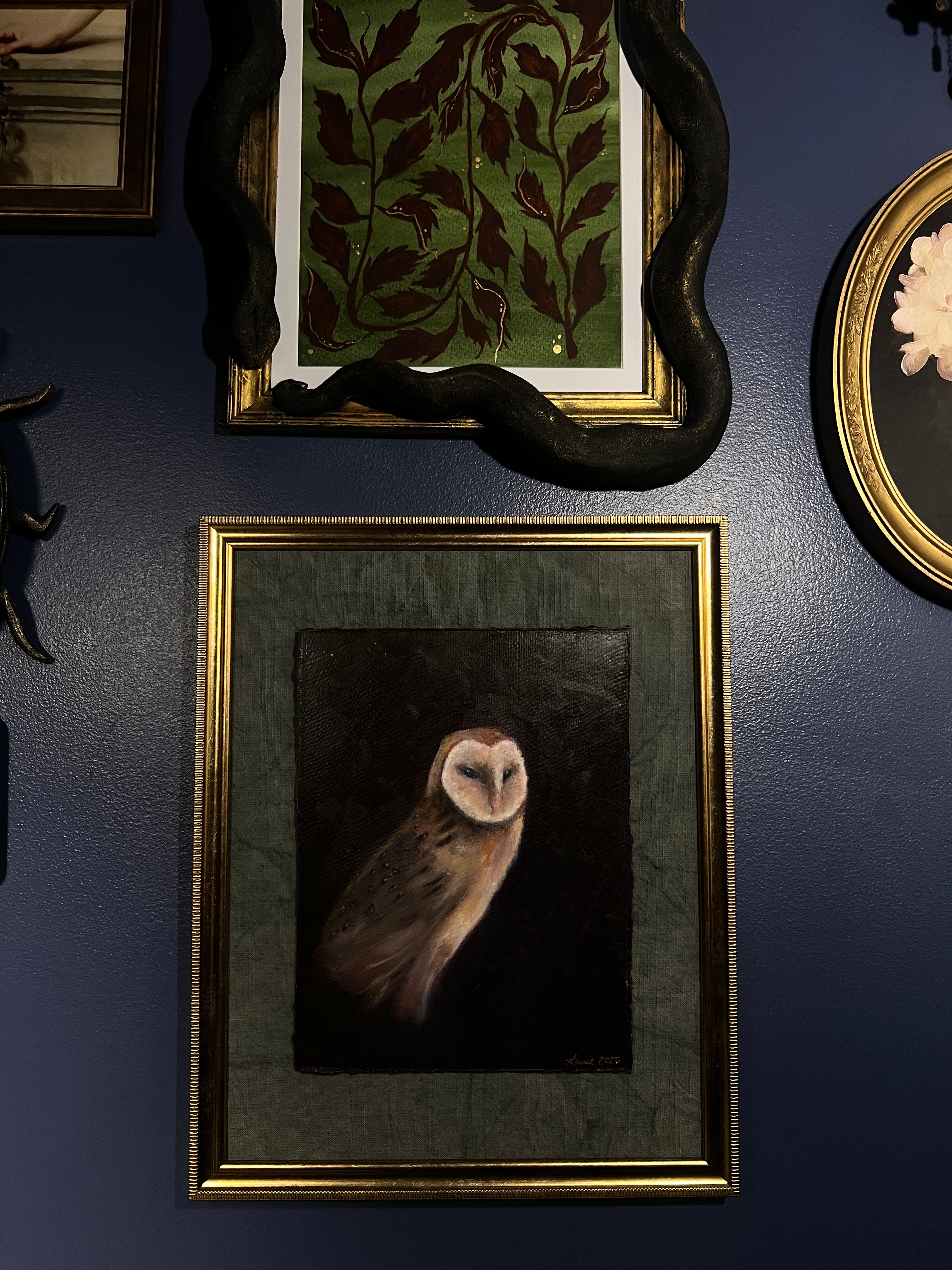 A framed art print depicting a barn owl on a dark background, mounted on a dark blue textured wall alongside other framed pieces.