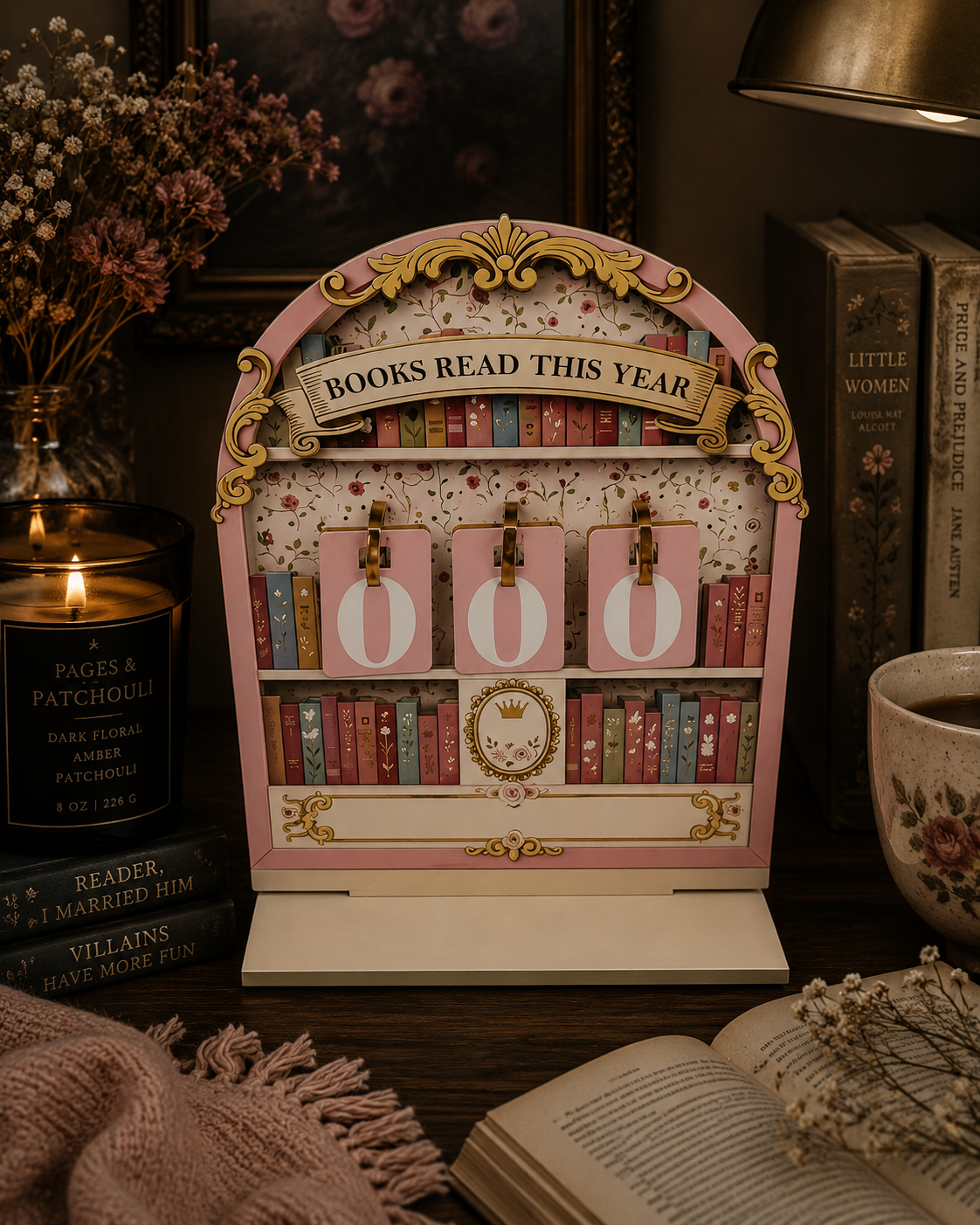 A decorative pink and gold book-themed year counter display reading 'BOOKS READ THIS YEAR 2025', styled on a dark wooden table with books, a candle, and dried flowers in a cozy room setting.