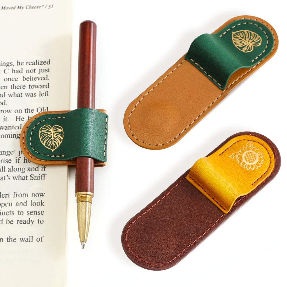 Three leather bookmark holders with metal clip tabs are arranged on a white background, alongside a brown pen resting in one holder and an open book page on the left. The holders feature stitched edges, gold-tone leaf and sunflower motifs, and different color combinations: dark green with tan, mustard yellow with brown, and dark brown with yellow.