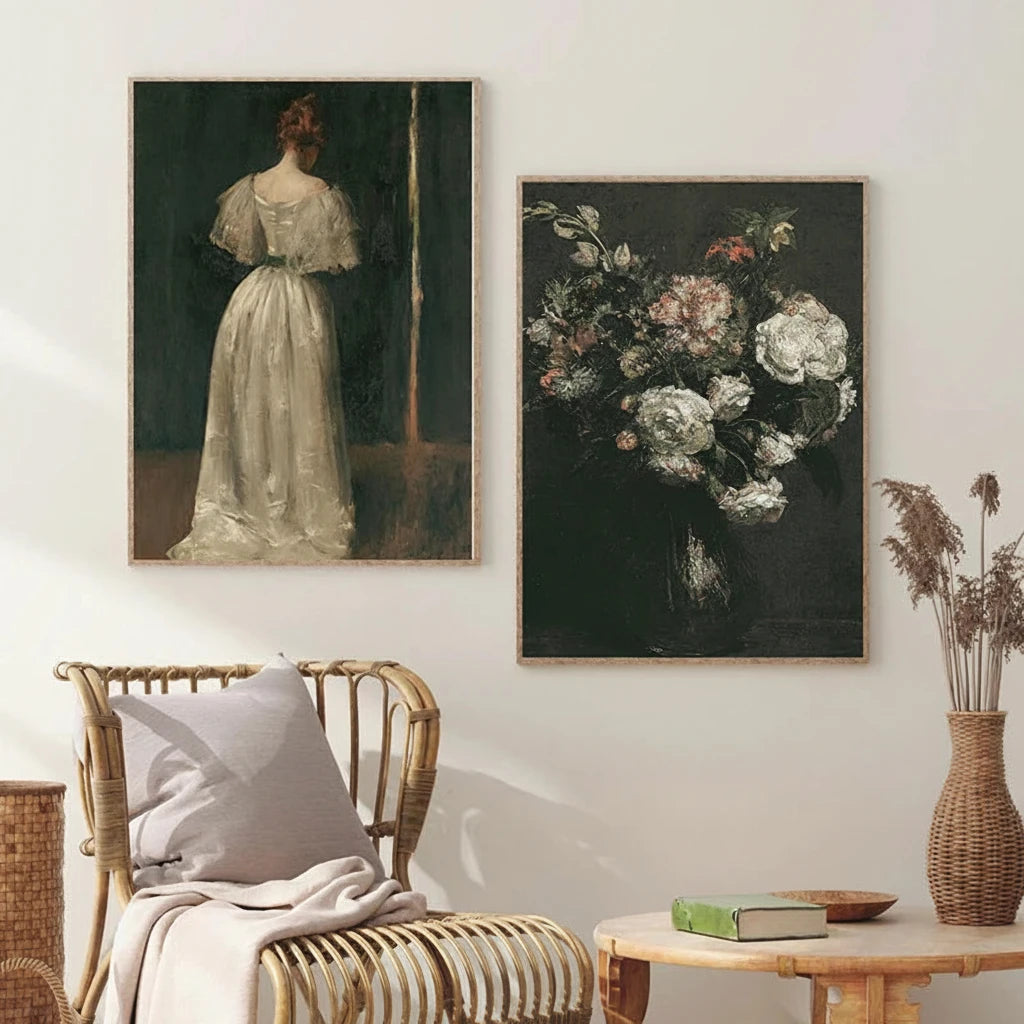 Two framed art prints displayed on a light beige wall above a rattan armchair and a small wooden side table. The left print shows a woman in a white dress from the back, and the right print shows a bouquet of white and pink flowers against a dark background.