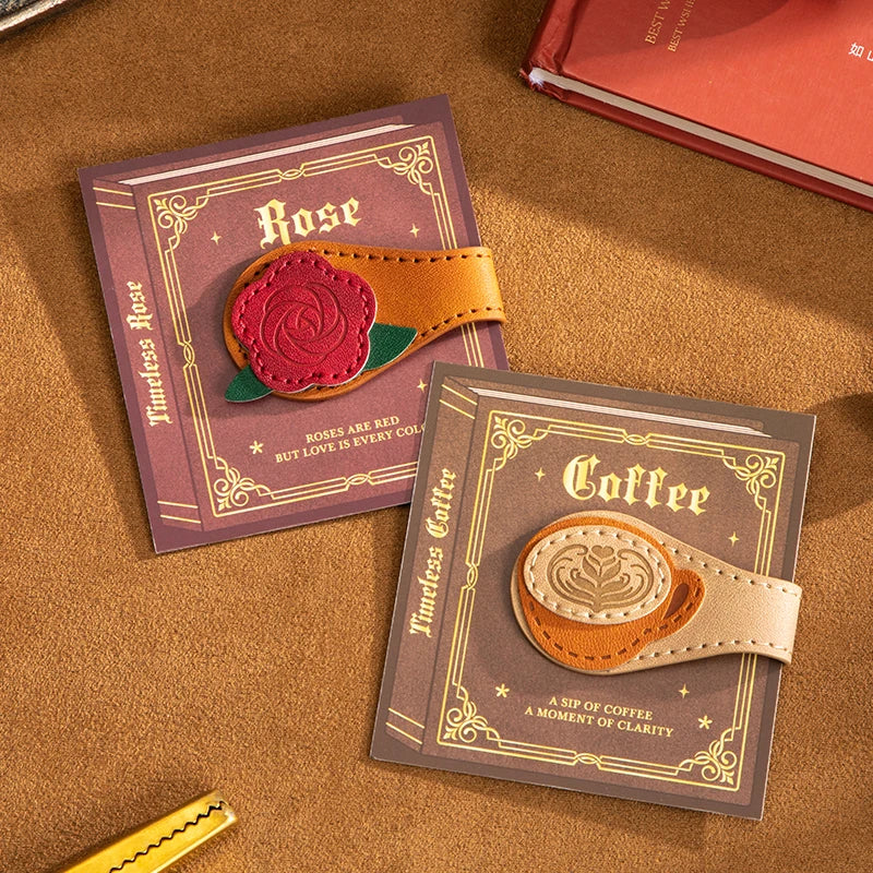 Two leather bookmark-style accessories displayed on decorative cards on a brown textured surface. One card is labeled 'Rose' with a pink rose-shaped leather piece attached to tan leather loops; the other card is labeled 'Coffee' with a beige coffee cup-shaped leather piece attached to tan loops.