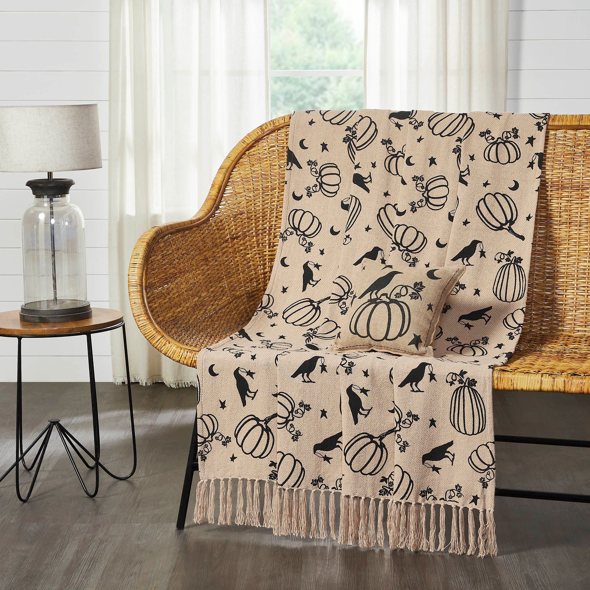 A beige throw blanket with a black pumpkin, crow, moon, and star pattern is draped over a wicker bench in a styled room setting.